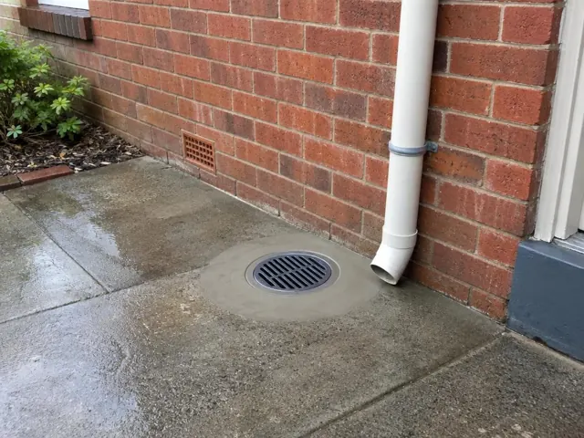 Clean outdoor drain grate beside brick wall and downpipe on wet concrete paving in Lambton